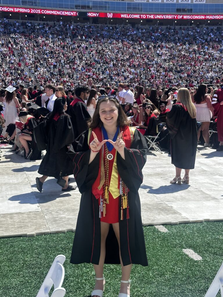 Elizabeth Preboski stands in Camp Randall wearing a undergraduate graduation gown making the letter "W" with her hands.